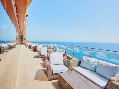 Terrassenbereich mit Rattanmöbeln und weißen Polstern, Balkon mit Glasgeländer, Blick aufs blaue Meer.