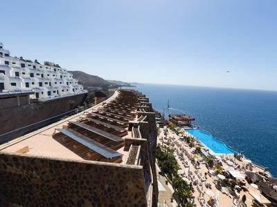 Terrassenförmige Anlage mit weißer Gebäudezeile, Blick auf blauen Ozean und klaren Himmel.