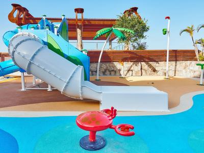Wasserpark mit blauer Rutsche, kleinem Pool, roten Spielgeräten und Palmen im Hintergrund.