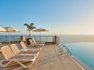 Piscine à débordement avec chaises longues et vue sur la mer au coucher du soleil.