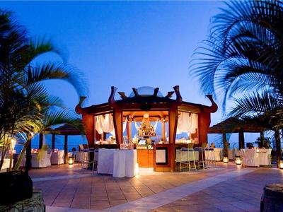 Abendliche Gartenbar mit warmem Licht, Palmen und Holzmöbeln unter klarem blauem Himmel.