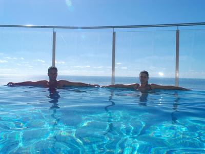 Zwei Personen liegen entspannt in einem klaren, blauen Pool mit Glaszaun und Meerblick im Hintergrund.