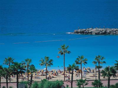 Strand mit Palmen, Sonne, blauem Meer, zwei Segelbooten und felsigem Wellenbrecher im Hintergrund.