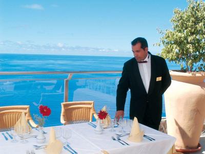 Mann in schwarzem Anzug richtet weißen Tisch mit roten Rosen und Meerblick bei Sonnenschirm her.