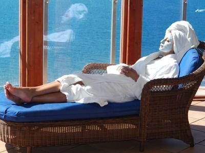 Personne se détend en peignoir blanc sur une chaise longue en osier avec coussins bleus, vue sur la mer en arrière-plan.