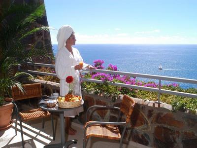 Frau in weißem Bademantel auf Balkon mit Blumen, Blick aufs blaue Meer und klaren Himmel.
