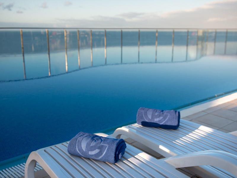 Moderner Infinity-Pool mit Glasgeländer und zwei weißen Liegestühlen mit blauen Handtüchern.