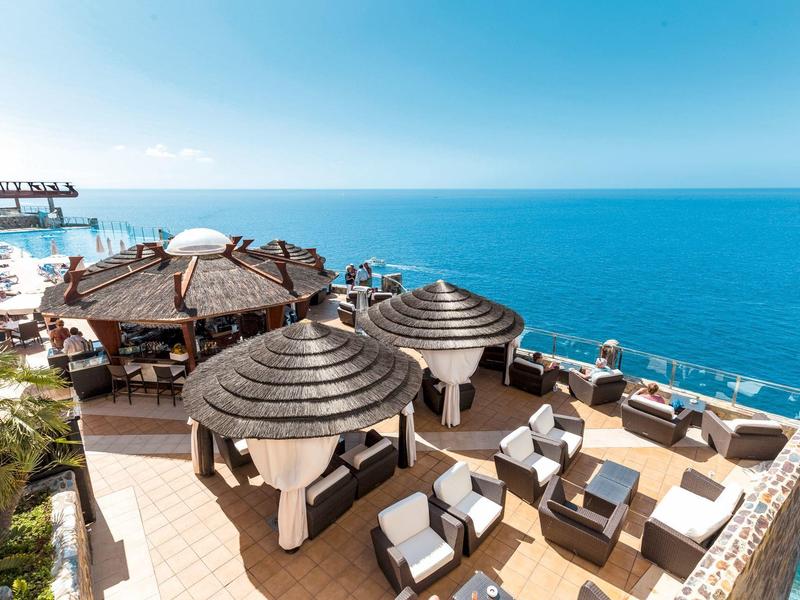 Terrasse mit runden Strohdächern, weißen Sesseln und Blick auf ruhiges blaues Meer.