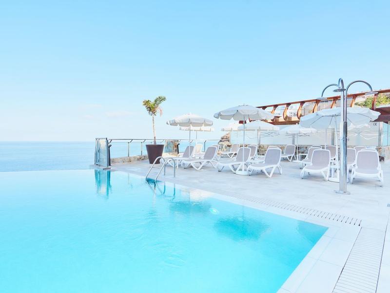 Infinity-Pool mit klarblauem Wasser und weißen Liegestühlen unter Sonnenschirmen bei klarem Himmel.