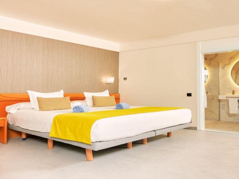 Modernes Hotelzimmer mit Doppelbett, gelben und weißen Akzenten, Sitzbereich und sichtbarem Bad.