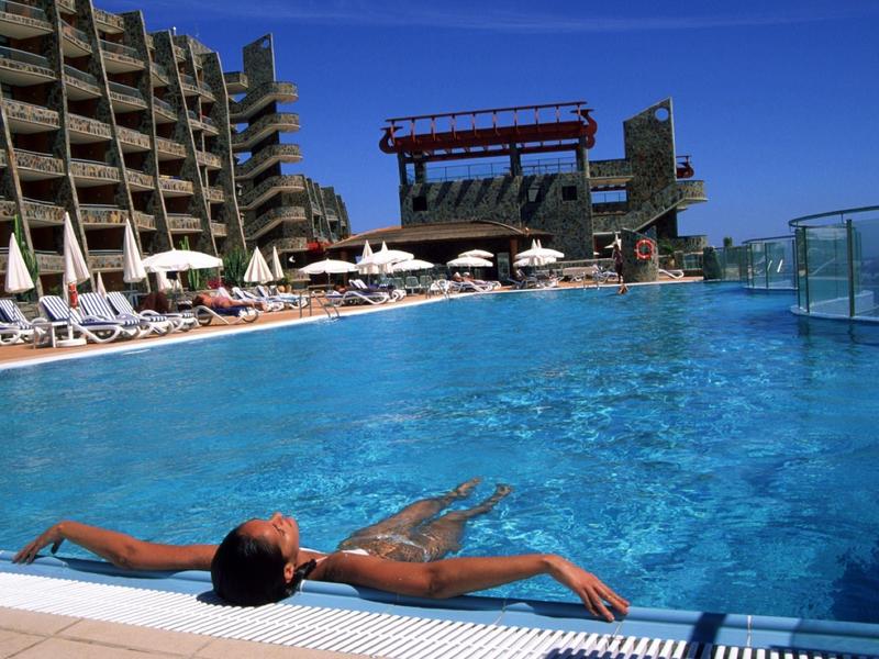 Großes Außenpool mit klarblauem Wasser, umgeben von Liegestühlen und Sonnenschirmen, Gebäude im Hintergrund.