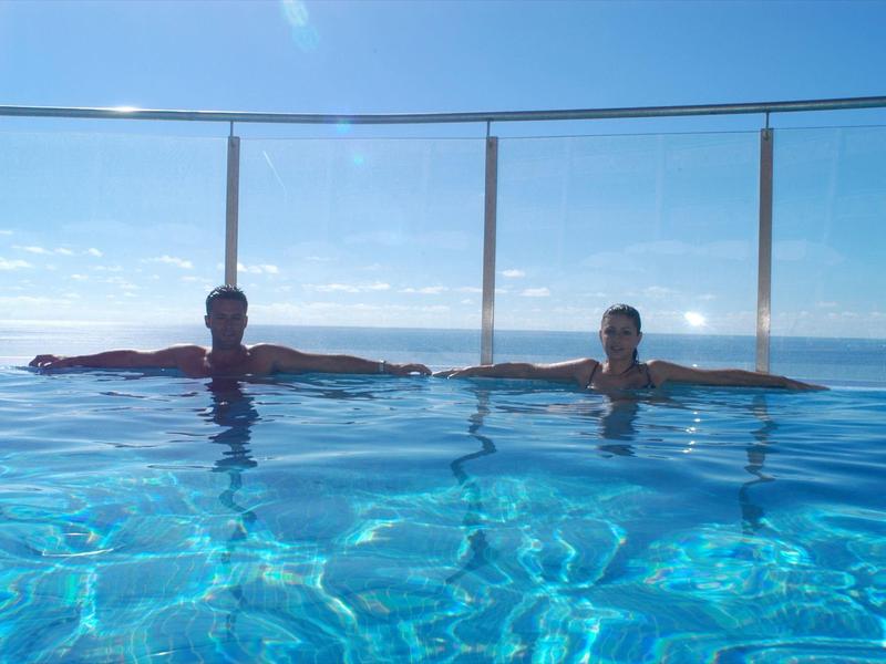 Zwei Personen liegen entspannt in einem klaren, blauen Pool mit Glaszaun und Meerblick im Hintergrund.