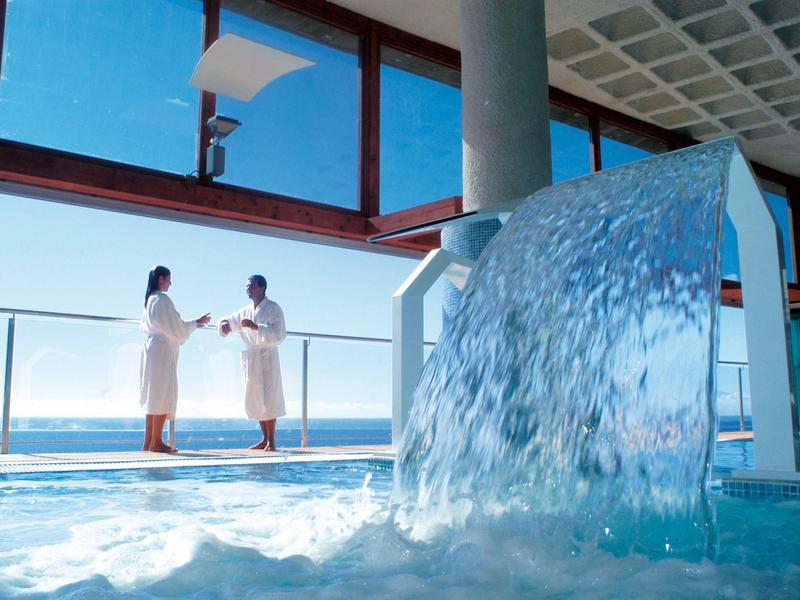 Innenpool mit Wasserfall, großer Fensterfront und zwei Personen in weißen Bademänteln.