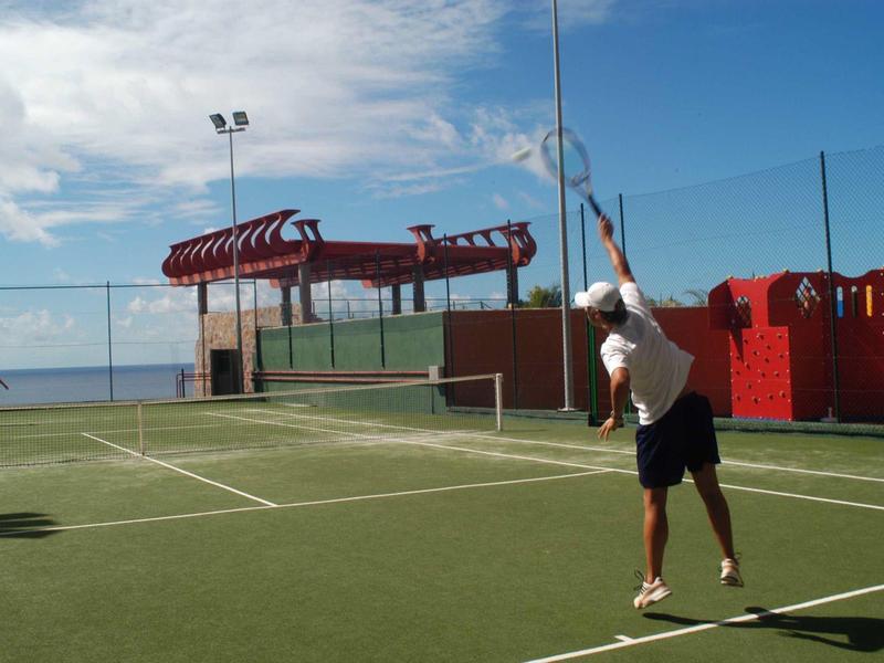 Zwei Personen spielen Tennis auf einem Außenplatz mit grünem Belag und blauem Himmel.