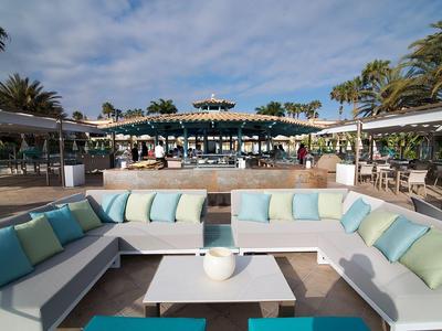 Espace lounge moderne avec canapés blancs, coussins clairs et palmiers près de la piscine de l'hôtel.