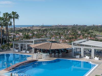 Piscine d'hôtel avec bar et palmiers, vue sur la ville côtière et la mer en arrière-plan.