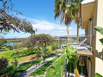 Balcon donnant sur des plantes tropicales, un chemin de jardin et une eau bleue sous un ciel dégagé.
