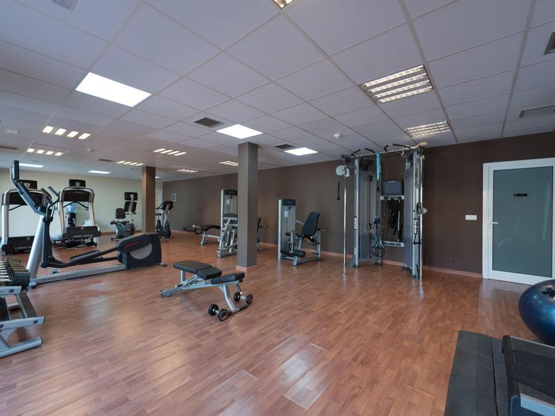 Salle de sport lumineuse avec sol en bois, divers appareils de musculation et grands miroirs au mur.