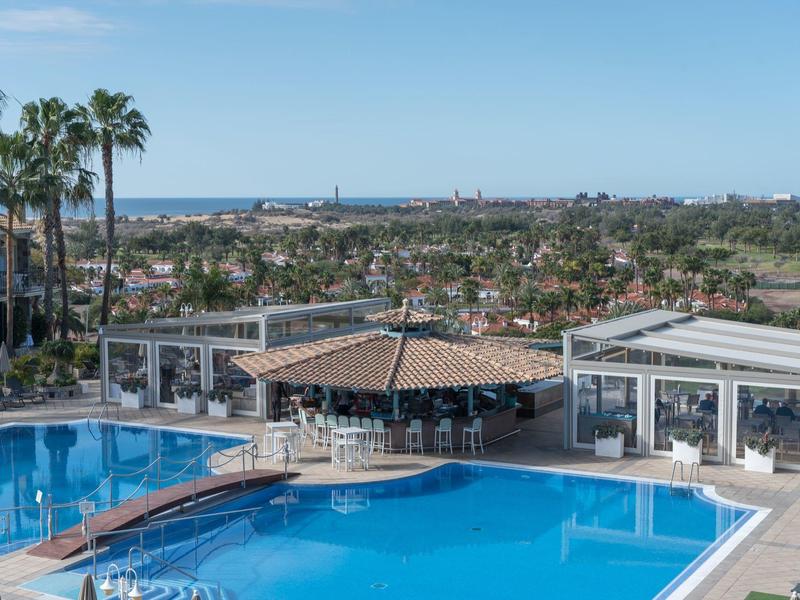 Piscine d'hôtel avec bar et palmiers, vue sur la ville côtière et la mer en arrière-plan.