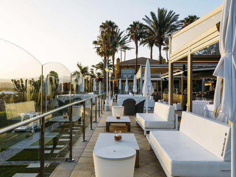 Terrasse avec canapés blancs et tables en plein air avec vue sur des palmiers et le coucher de soleil.