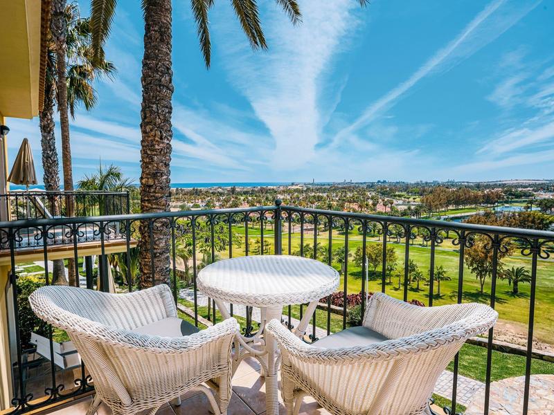 Balcon avec chaises blanches et table surplombant un terrain de golf et un paysage urbain sous un ciel bleu.