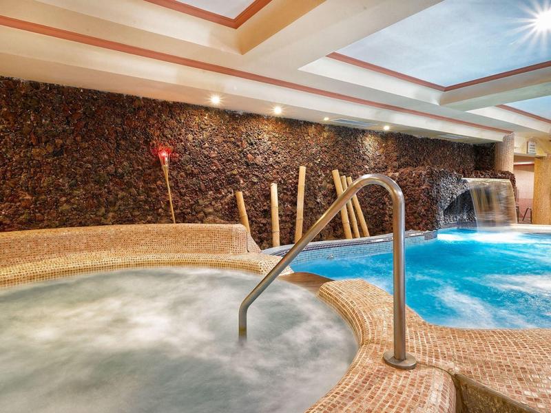 Piscine intérieure avec jacuzzi et cascade, éclairage chaleureux et mur en pierre naturelle.