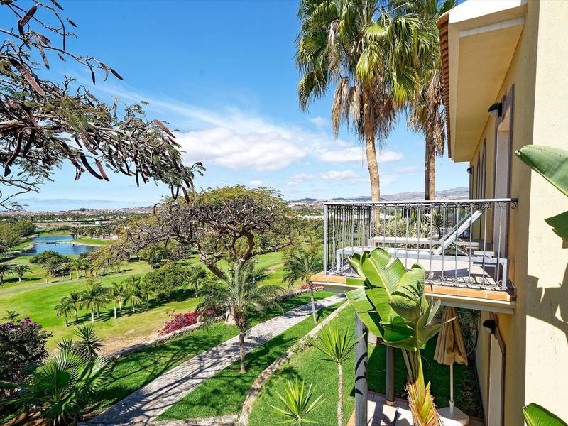 Balcon donnant sur des plantes tropicales, un chemin de jardin et une eau bleue sous un ciel dégagé.