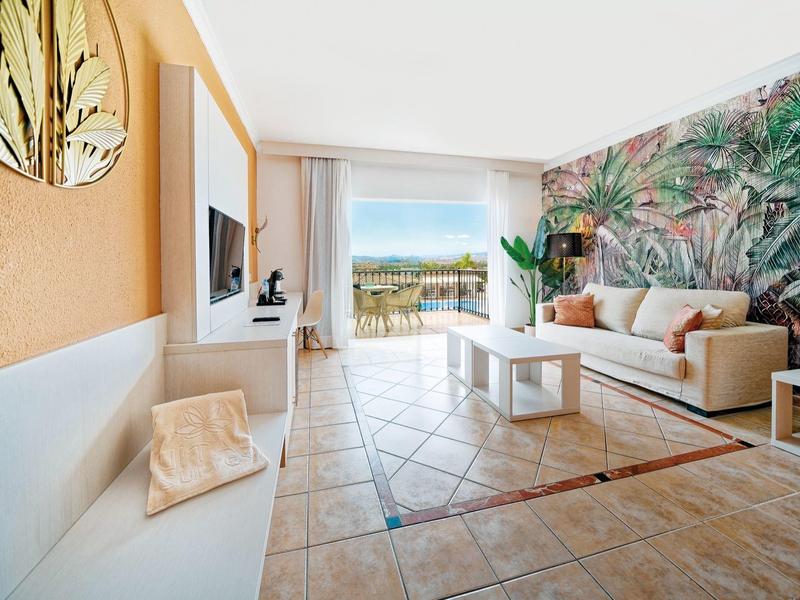 Salon lumineux avec décoration murale tropicale et accès à une terrasse avec vue sur la mer.