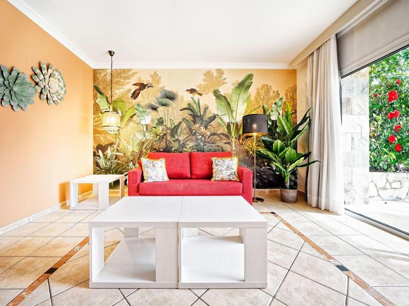 Salon lumineux avec canapé rouge, décoration murale tropicale et grandes fenêtres donnant sur le jardin.