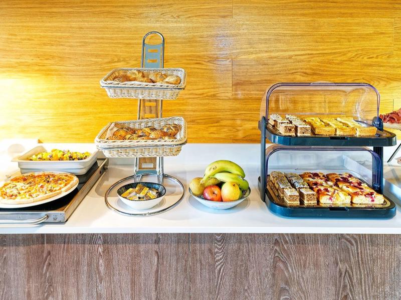 Buffet avec diverses pâtisseries, fruits et pizza sur un comptoir blanc devant un mur en bois.