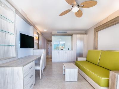 Salon lumineux avec canapé vert, bureau blanc et ventilateur au plafond.