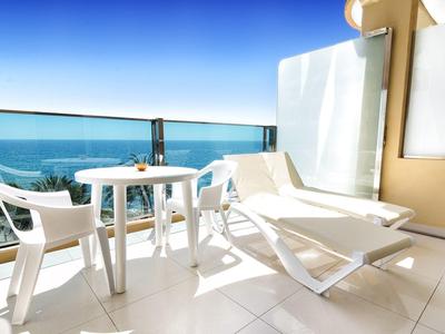 Balcon d'hôtel avec chaise longue blanche, table et chaises surplombant la mer bleue.