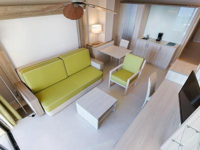 Salon moderne avec canapé vert, fauteuil, table basse et télé dans un intérieur lumineux.