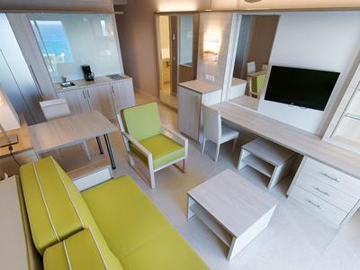 Salon d'hôtel moderne avec canapé vert, fauteuil, table basse et grande télévision.