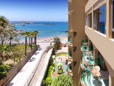 Vue depuis un balcon d'hôtel sur une plage ensoleillée avec palmiers et océan bleu.