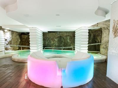 Espace spa luxueux avec jacuzzi, sièges éclairés en dégradé de rose et bleu, murs en pierre.
