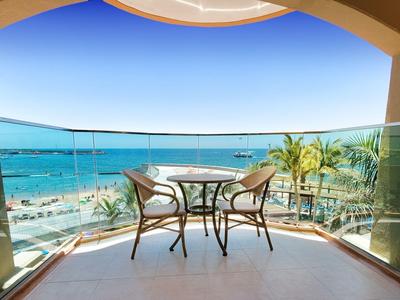 Balkon mit Glasgeländer, zwei Stühlen und Tisch mit Meerblick und Strand im Hintergrund
