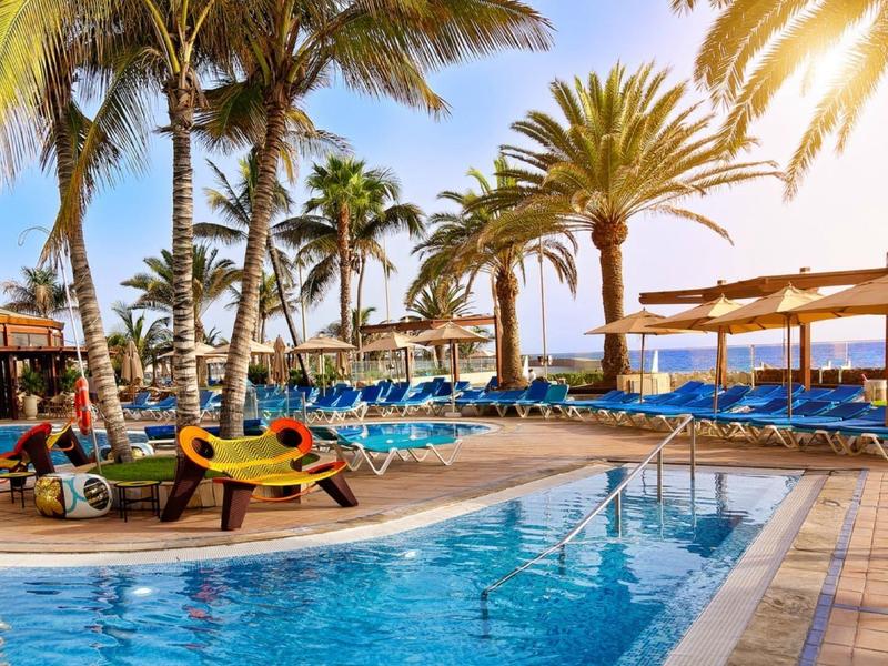 Piscine extérieure avec transats, parasols et palmiers sous un ciel ensoleillé.