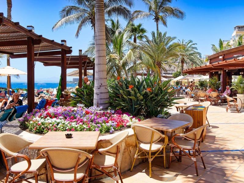 Terrasse d'un restaurant avec vue sur la plage, palmiers et chaises en rotin sous pergola.