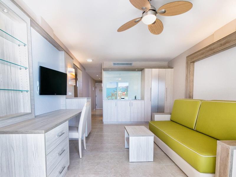 Salon lumineux avec canapé vert, bureau blanc et ventilateur au plafond.