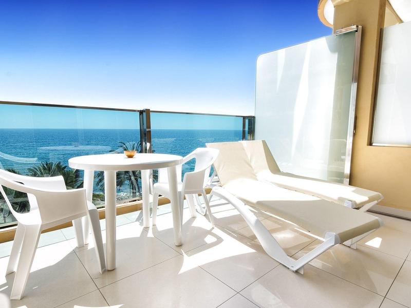 Balcon d'hôtel avec chaise longue blanche, table et chaises surplombant la mer bleue.