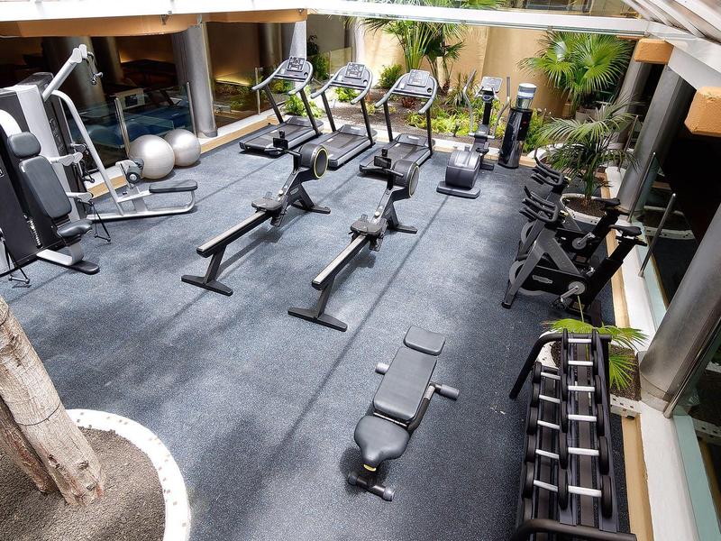 Salle de sport bien équipée avec rameurs, poids libres, et machines dans un espace lumineux.