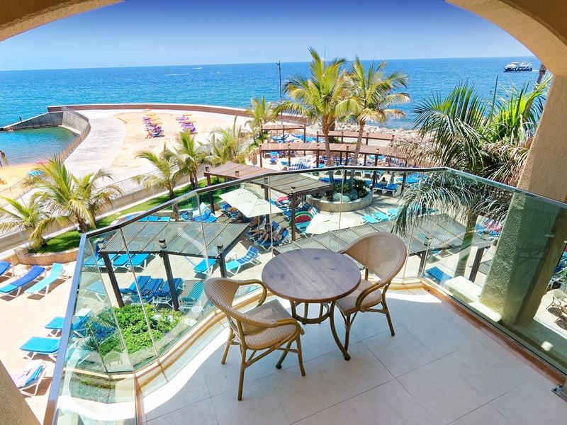 Balcon d'hôtel avec table et chaises donnant sur une plage sableuse et une mer bleue.