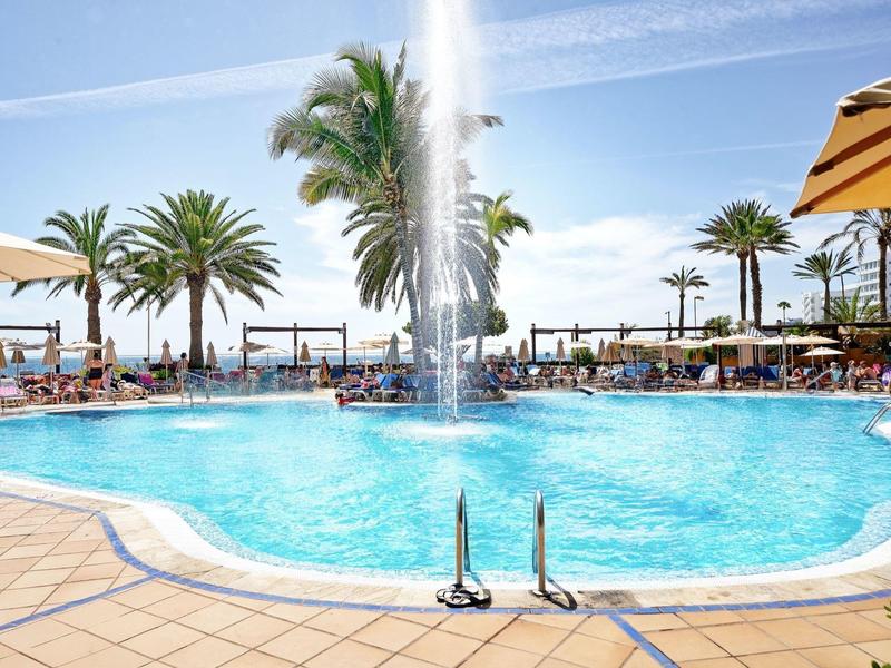 Piscine extérieure entourée de palmiers et de chaises longues sous un ciel bleu.