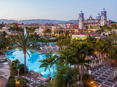 Hotelanlage mit großem Pool, Palmen und umliegenden historischen Gebäuden bei Sonnenuntergang.