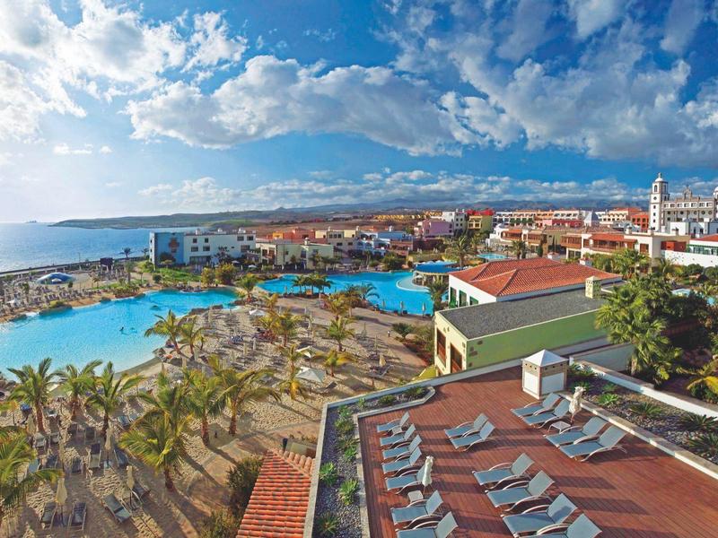 Vista de un hotel frente al mar con piscinas, palmeras y ciudad bajo cielo azul.