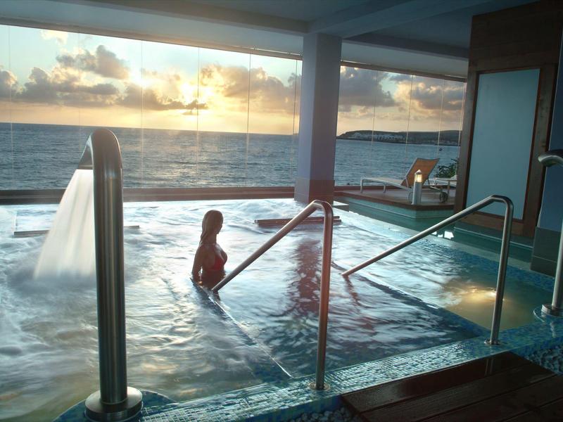 Persona relajándose en una piscina interior con vista al atardecer sobre el mar.