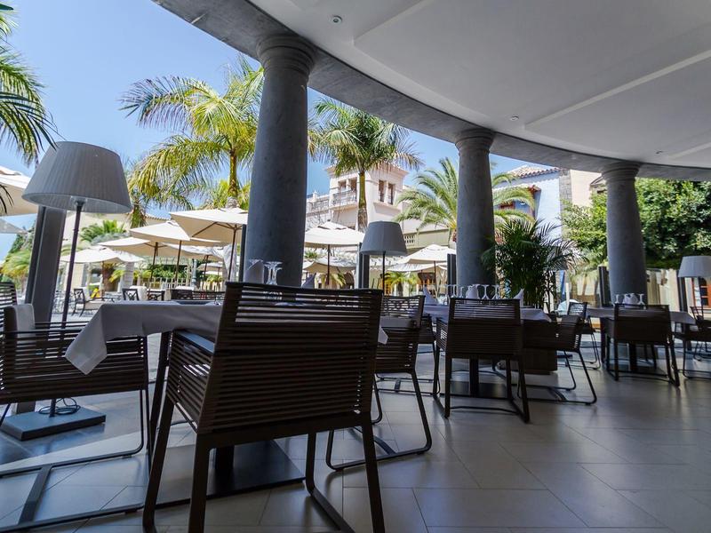 Área exterior moderna de un hotel con sillas, mesas y sombrillas bajo el sol.