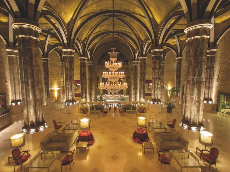 Área de lobby de hotel lujoso con candelabros y zonas de asientos en un gran salón abovedado.