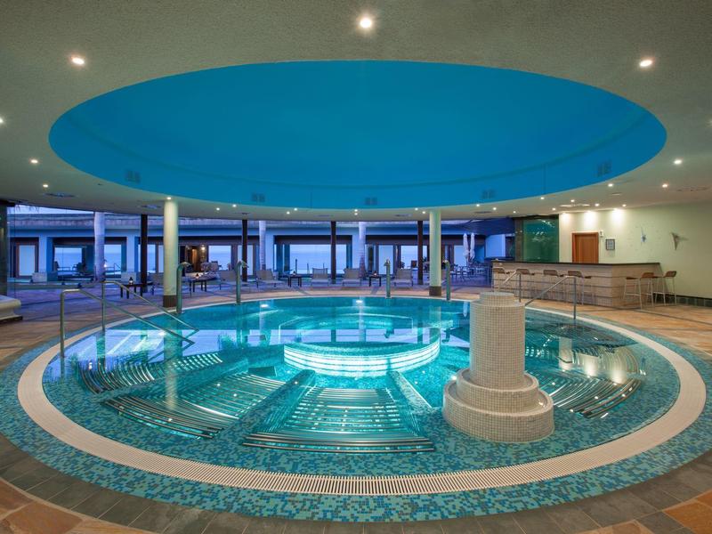 Piscina interior iluminada con jacuzzi circular y ventanas panorámicas en un hotel.